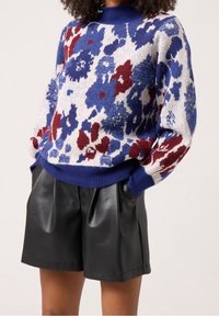 Pletený svetr v modré, červené a krémové barvě s květinovým vzorem, žebrovaným výstřihem a volným střihem, kombinovaný s vysokým pasem černých kožených šortek.