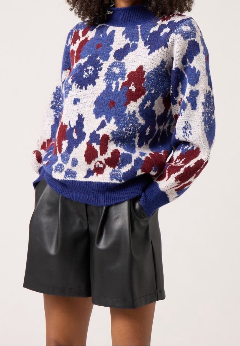 Pletený svetr v modré, červené a krémové barvě s květinovým vzorem, žebrovaným výstřihem a volným střihem, kombinovaný s vysokým pasem černých kožených šortek.