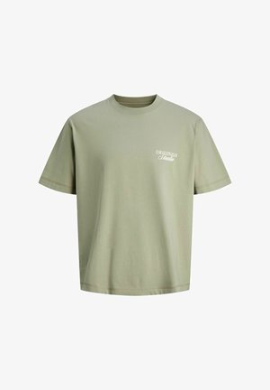 Ragazzo sorridente, con berretto bianco, giacca nera con zip sopra una maglietta verde, pantaloni neri larghi e scarpe da ginnastica bianche, in piedi su sfondo bianco.