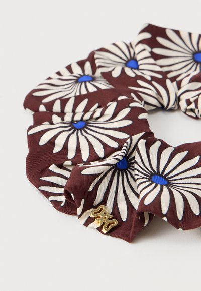 Chouchou en tissu marron avec des motifs de marguerites blanches aux centres bleus et un petit charme décoratif doré sur un fond blanc.