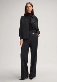 Zwarte op maat gemaakte outfit bestaande uit een button-up shirt met twee borstzakken, een geribbelde coltrui en brede broekspijpen met een gespsluiting.