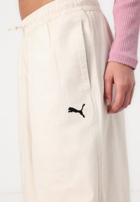 Ljust krämfärgade joggerbyxor i mjukt tyg, med ett elastiskt midjeband, sidofickor och en svart Puma-logotyp på vänster ben.
