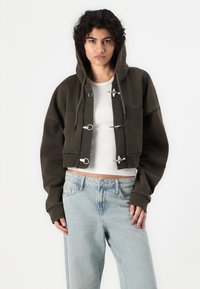 Cropped olive green hoodie with a drawstring hood, silver hardware closures, and large front pockets, paired with light blue jeans.