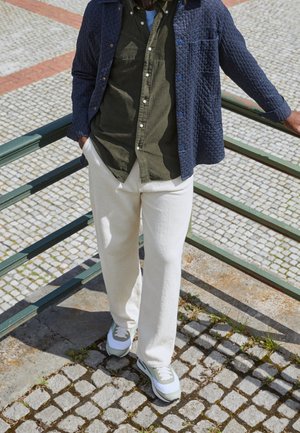 Granatowa pikowana kurtka na oliwkowej zielonej koszuli, w zestawieniu z kremowymi spodniami i szarymi trampkami. Stojący na brukowanym chodniku obok poręczy.