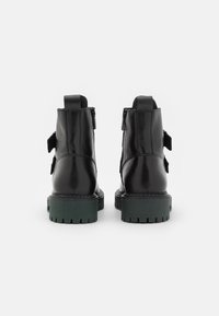Paire de bottines en cuir noir avec semelles épaisses vertes, languettes à l'arrière, fermetures éclair latérales et sangles décoratives sur fond blanc.