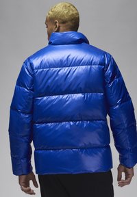 Blå pufferjakke med en blank overflade, der har horisontale quiltede mønstre og en høj krave. Designet er oversized og afslappet.