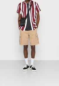 Kurzärmliges Hemd mit roten und weißen Streifen, darunter ein schwarzes T-Shirt, beigefarbene Cargo-Shorts, weiße Socken und schwarze Sneaker.