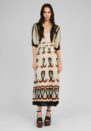 Femme aux longs cheveux foncés portant une robe midi à motifs crème, noire et orange avec des manches mi-longues et des sandales compensées noires, debout sur un fond gris.