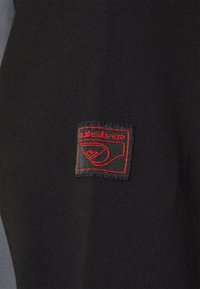 Svart sweatshirt med en tygpatch som har en röd "Quiksilver"-logotyp och designelement, som visar en slät textur och en atletisk stil.