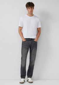 Weißes Baumwoll-T-Shirt mit kurzen Ärmeln, kombiniert mit dunkelgrauen Jeans. Das Model trägt weiße Sneakers mit braunen Akzenten.