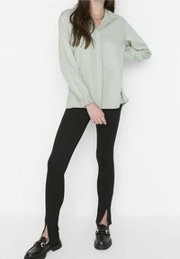 Light green, long-sleeve button-up shirt paired with black leggings featuring side slits, worn with black loafers. Smooth fabric, fitted design.