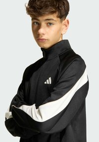 Čierna športová bunda s vysokým golierom, biele akcenty na rukávoch a reflexným logom na hrudi. Hladká tkanina so štruktúrovaným dizajnom.