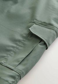 Gros plan sur une manche en tissu vert olive avec une poche à rabat rectangulaire et des coutures apparentes.