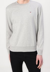 Osoba wearing a jasnoszary długi rękaw sweatshirt z małym czerwonym, białym i czarnym logo na piersi oraz czarnymi spodniami.