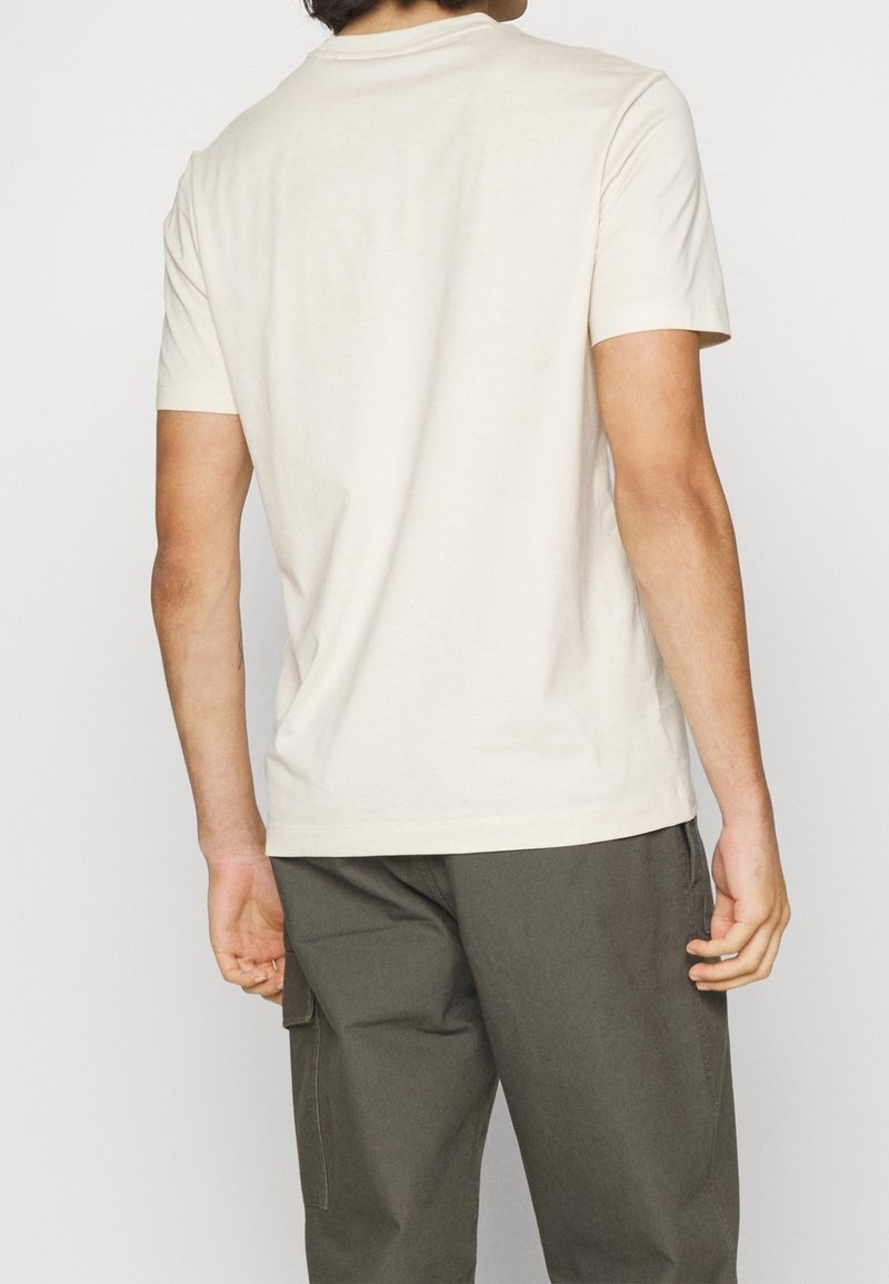 Homme portant un t-shirt uni à manches courtes blanc cassé et un pantalon cargo vert foncé, vu de dos sur fond blanc.