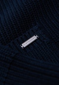 Tessuto lavorato a maglia blu navy con un motivo testurizzato, caratterizzato da un'etichetta argentata con scritto "QUINTESSENC" fissata da due rivetti.