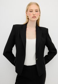 Blazer nero con spalle strutturate, revers a punta e un solo bottone dorato. Indossato sopra una canotta bianca a coste.