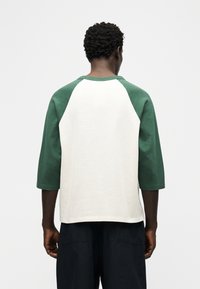 Άνδρας που φαίνεται από πίσω, φοράει λευκό πουκάμισο με πράσινα μανίκια ράγκλαν και μαύρο παντελόνι πάνω σε απλό ανοιχτό φόντο.