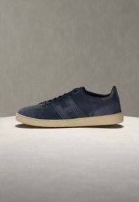 Baskets en daim bleu marine avec semelle beige, design à lacets, et un grand "H" sur le côté, présentées sur un fond neutre.