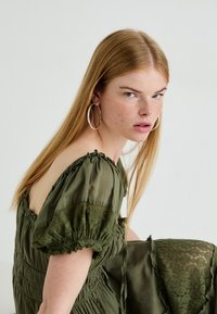 Olijfgroene jurk met poffende mouwen, smock- manchetten en kantaccenten, gestyled met een relaxte snit en zijstrikken.