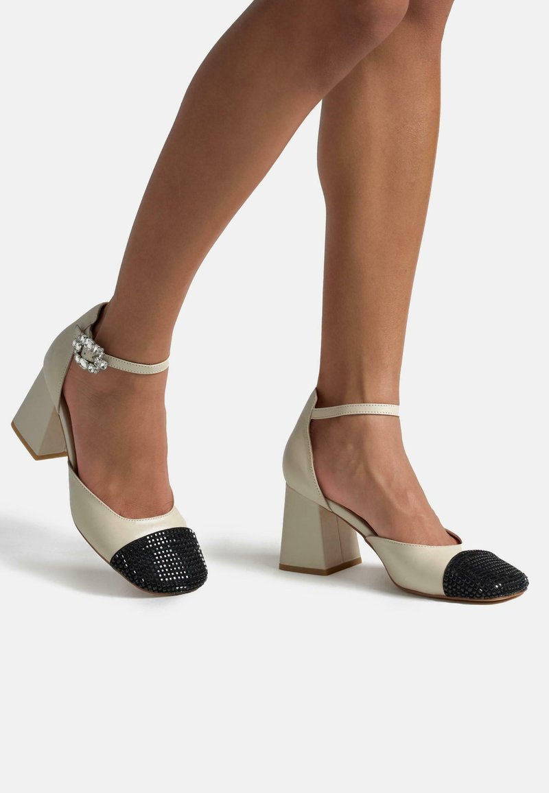 Scarpe in pelle beige con punta in strass neri, tacco a blocco e cinturino alla caviglia con una chiusura decorativa. Texture morbida in generale.