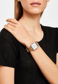 Montre carrée avec un boîtier en ton argent et un bracelet en cuir brun, portée au poignet. Indicateurs d'heure et aiguilles simples sur un cadran blanc.