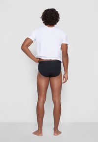 Schwarze Schwimmslips mit einer glatten Textur und einem flachen Bund. Getragen unter einem einfach weißen, lässig geschnittenen T-Shirt, von hinten betrachtet.
