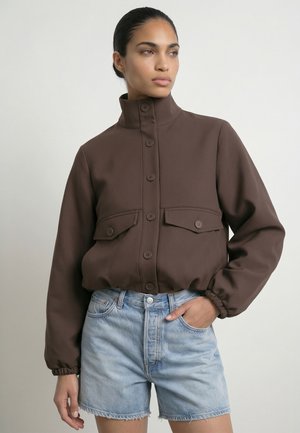 Kvinde iført en brun jakke med høj krave, knapper og klaplommer samt lyseblå denimshorts, stående med den ene hånd i lommen.