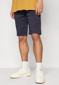 Shorts in cotone blu con tasche laterali e taglio dritto, abbinati a sneaker bianche e calzini bianchi a coste. Design semplice, senza motivi visibili.