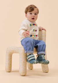 Maglione a maglia crema con testo colorato, pantaloni di denim blu e scarpe con velcro verdi, seduto su una sedia a trama, semi-trasparente e maculata.