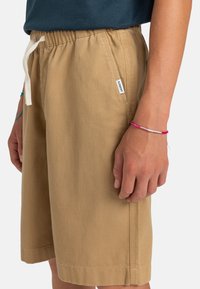 Shorts en coton beige avec une taille élastique, cordon de serrage réglable et poches sur les côtés. Présente une petite étiquette de marque sur le côté.