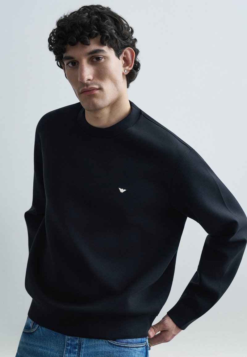 Čierna mikina s vysokým golierom, s malým strieborným logom na hrudi. Tkanina pôsobí hladko s voľným strihom. Viditeľné džínsové rifle.