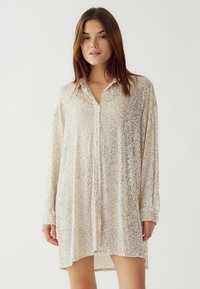 Jauna moteris vilki ilgomis rankovėmis, su sagomis, šviesiai ruda blizgučių suknelė, stovi prieš paprastą šviesų foną.