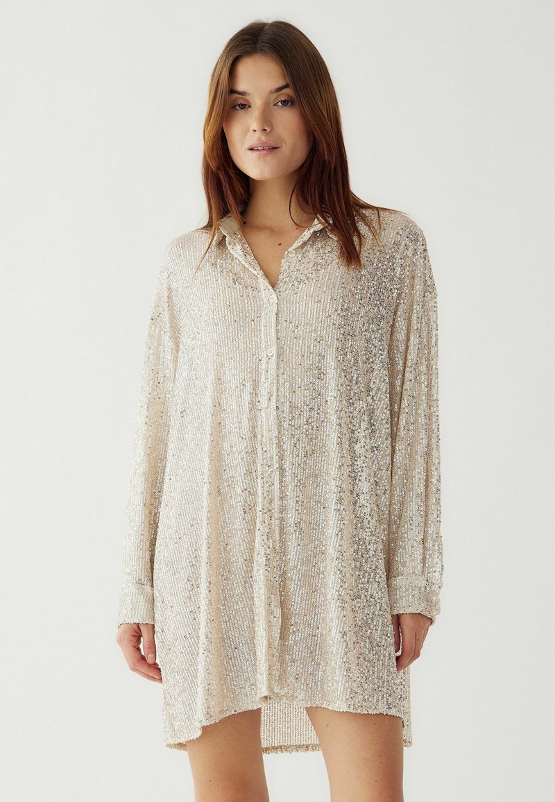 Jauna moteris vilki ilgomis rankovėmis, su sagomis, šviesiai ruda blizgučių suknelė, stovi prieš paprastą šviesų foną.