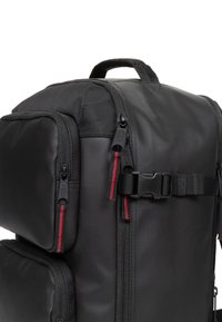 Mochila negra hecha de material duradero, con múltiples compartimentos con cremallera y cremalleras de acento rojo, con un asa superior y una correa ajustable.
