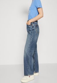 Person som bär en blå grafisk t-shirt, hög midja avslappnade blå jeans och vita plattformssneakers står i profil.