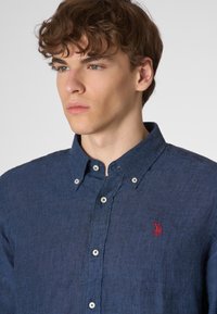 Camicia di lino blu navy con colletto button-down, bottoni bianchi e un piccolo logo rosso sul lato sinistro del petto. Dettaglio in tessuto testurizzato visibile.
