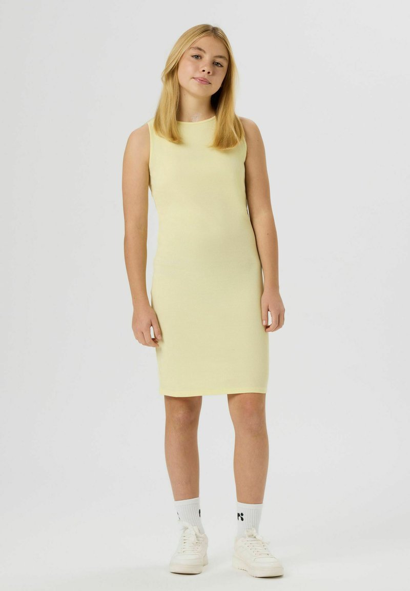 Jeune femme debout portant une robe sans manches jaune pâle jusqu'aux genoux, des chaussettes blanches et des baskets blanches sur un fond clair uni.