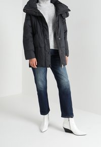 Černá péřová bunda s vysokým límcem a kapucí, nošená přes šedý rolák, doplněná modrými džínami a bílými kotníkovými botami.