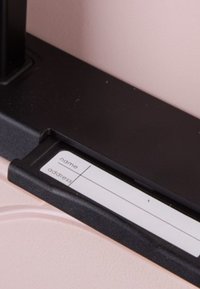 Manico in plastica nera con un portalabel che presenta spazi vuoti per "nome" e "indirizzo", attaccato a una superficie rosa chiaro.