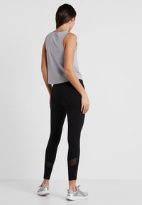 Szary krótki top na ramiączkach w zestawieniu z czarnymi legginsami z siateczkowymi wstawkami na łydkach. Model ma na sobie srebrne buty sportowe. Gładkie tekstury w całym zestawie.