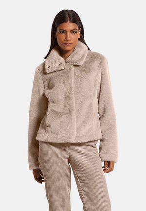 Frau trägt eine beige Kunstpelzjacke mit weitem Kragen und beige gerippte Cordhosen, steht vor einem schlichten Hintergrund.
