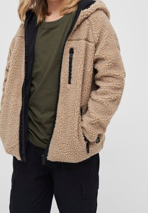 Beige Fleecejacke mit Kapuze, Reißverschluss vorne und zwei Seitentaschen. Schwarze Akzente und ein strukturiertes, flauschiges Gewebe. Kombiniert mit einem grünen Oberteil.