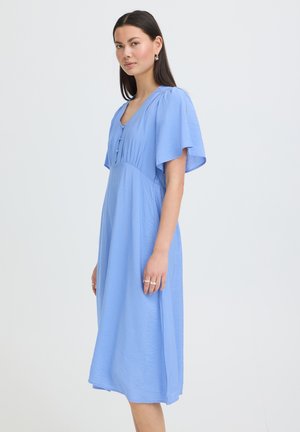 Ung kvinde med glat mørkt hår iført lyseblå mellemlang kjole med flagrende ærmer, stående sidelæns mod en ensfarvet hvid baggrund.