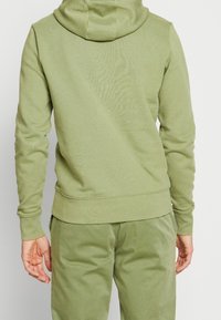 Felpa color verde oliva realizzata in tessuto morbido, con cappuccio regolabile, maniche lunghe e orlo a coste. Indossata con pantaloni verdi abbinati.
