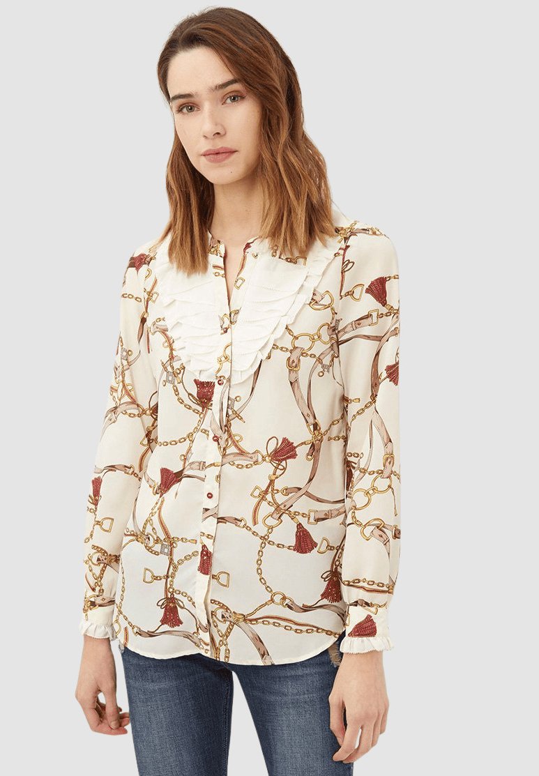 Blusa color crema con un cuello con volantes, que presenta un patrón de cadenas y borlas en rojo y dorado, mangas largas y cierre de botones.