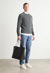 Maglione grigio a maglia, camicia bianca sotto, jeans blu, tiene una borsa a tracolla in pelle nera con il logo "HUGO" in rilievo; sneakers bianche.
