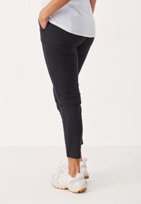 Person som bär svarta slim-fit byxor och vita sneakers med beige sulor, står med händerna i fickorna mot en enkel ljus bakgrund.