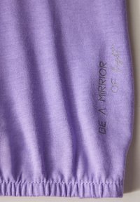 Lila Stoff mit glatter Textur. Verfügt über elastische Bündchen und den Text "SEI EIN KRIEGER DES Lichts" in einer kontrastierenden silbernen Farbe.