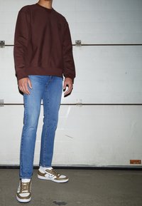 Brun sweatshirt, ljusblå jeans och bruna och vita sneakers. Slätt tyg, figursydd design, kontrasterande färger och synliga sömnadsdetaljer.